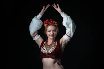 woman in traditional folk eastern slavic costume for dance with circlet of flowers, vest, sleeves and necklace looking at camera and holding hands over head isolated on black
