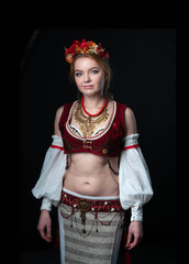 beautiful woman in traditional folk slavic costume for belly dance and trible with circlet of flowers, vest, skirt, sleeves and necklace standing and looking at camera on black background