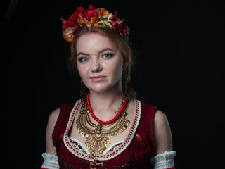 portrait of attractive woman in traditional costume in ukranian national stile with circlet of flowers, red vest and golden necklace looking at camera isolated on black