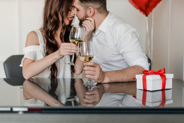 happy handsome man and woman drink white wine at home with present in gift box and red heart shaped balloons at home