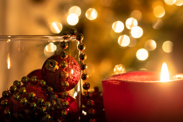 Christmas candle close-up view with glowing flame and ornaments in a crystal jar with blurred tree lights as background.