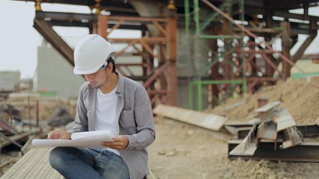 Asian construction engineer worker with protective helmet and looking blueprints paper at building construction site