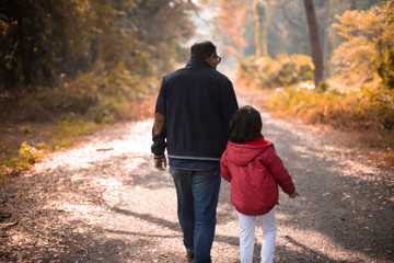 Back side of an Indian Bengali father and his little daughter with winter garments are walking through a pathway within a green natural forest in an afternoon. Indian lifestyle, fall and winter.