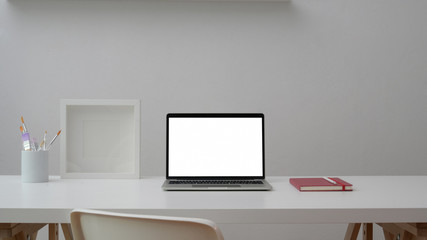 Cropped shot of workspace with open blank screen laptop, frame, notebook and painting brush on white table with white wall