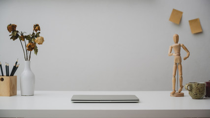Close up of workspace with laptop, pencils, wooden figure, dry roses vase and cup on white desk with white wall