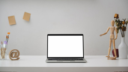 Close up view of  workspace with blank screen laptop, painting tools, wooden figure and decorations on white desk with white wall