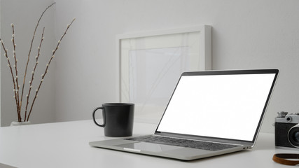 Cropped shot of  workspace with blank screen laptop, coffee cup, mock up frame and camera on withe desk with white wall