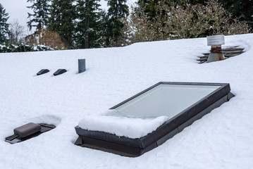 Residential house rooftop covered in fresh snow, skylight, roof vents, evergreen trees