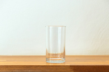 Empty glass of water on a wooden table and white wall with soft sunlight through a window.