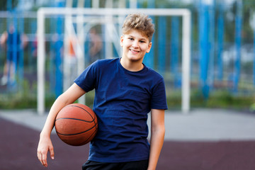 Cute boy in blue t shirt plays basketball on city playground. Active teen enjoying outdoor game with orange ball. Hobby, active lifestyle, sport for kids. 
