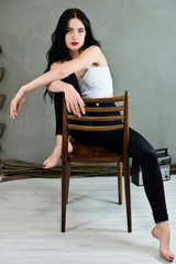 The concept of a glamorous female portrait. Portrait of a pretty brunette girl with long hair with excellent makeup in jeans and a T-shirt on a gray background sitting on a chair.