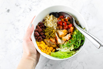 Mexican Lunch bowl in woman's hand on marble background. Salad bowl with corn, red beans, tonato with red onion, jalapenios, chicken breast, taco chips and yougurt garlic sauce. Top view