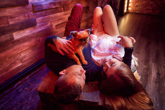 Beautiful Girl In White Shirt And Guy In Black Sweater With Small Dog On The Couch With Hay In The Barn Or In The Hayloft With Wooden Walls. Couple In Rustic Interior During Photo Shoot