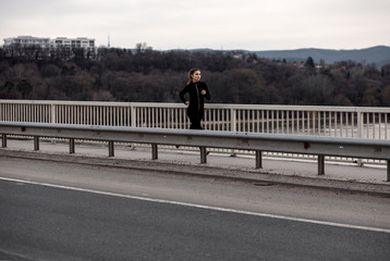 Young woman in black sports outfit running on the bridge in the city during day.