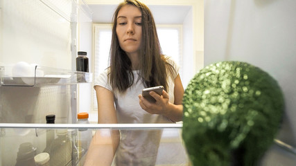 woman at fridge with smartphone making list of necessary food at home kitchen. Smart shopping list