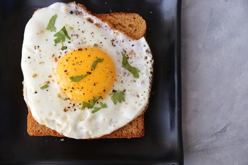 Fried Egg on Whole wheat Toast with salt and pepper for classic Breakfast. sunny side up egg with brown bread on black plate over wooden table, top view, copy space.