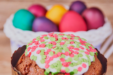 Easter composition. Easter cake on a stand made of a slice wood and a white basket on wheels with colorful Easter eggs on a wooden background. Close-up
