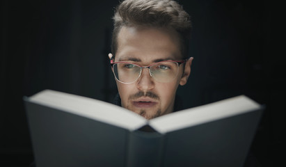 Close-up of serious young bearded man in glasses behind book isolated on black background