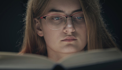 Close-up of concentrated girl in glasses behind book isolated on black background