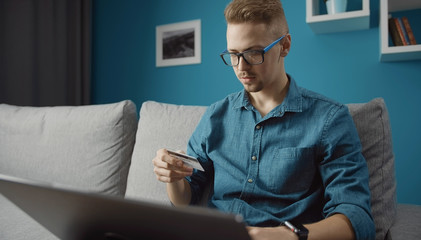 Casual young man doing online purchase by credit card using laptop staying home