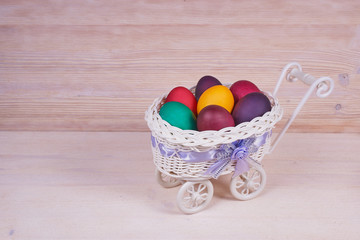 Easter composition. White basket on wheels with colorful Easter eggs on a wooden background. With copy space