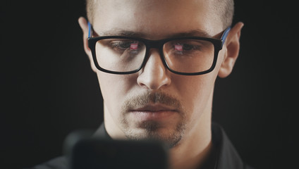 Close-up of attractive serious young man using smartphone, reflection of his hand and phone screen in his eyewear, isolated black background