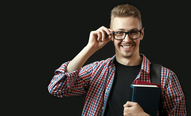 Portrait of delighted male student touching his eyeglasses and holding books confident in his knowledge isolated on dark blackboard and looking at camera