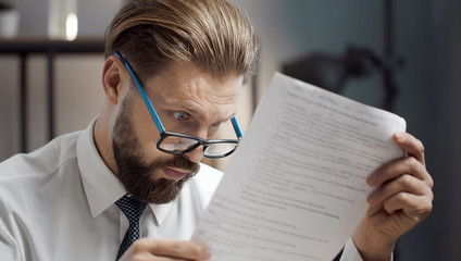 Close-up of astonished bearded businessman wearing glasses reading document with wide open eyes at...