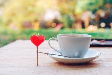 A red heart shaped candle with a cup of coffee and a coffee spoon on a saucer placed on a wooden table in the garden. Valentine day and love concept.