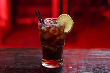 Closeup of a Cuba Libre cocktail in long glass, gin, standing on the bar counter, isolated on a red...