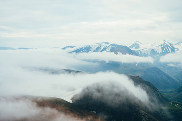 Obraz premium Beautiful atmospheric alpine landscape to big snowy mountain ridge with glacier. Giant low clouds among rockies in valley. Wonderful tranquil highland scenery. Flying over mountains above clouds.