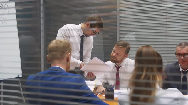 Young handsome caucasian businessman makes report presenting documents in front of conference table at meeting room, persuading investors. Shot through glass window of next office