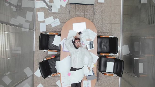Top view shot. Overworked male manager office worker lies on the table. Businessman start-up owner throws documents, papers in air. Indifferent business person going mad despair