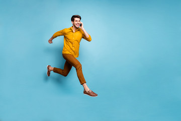 Fototapeta na wymiar I'm on my way. Full length photo of crazy guy jumping high rushing to party holding telephone speaking friends wear yellow shirt pants isolated blue color background