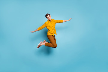 Full length photo of crazy guy jumping high holding hands spread by sides pretending bird flight wear yellow shirt trousers isolated blue color background