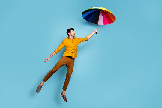 Full length photo of crazy guy jumping high holding colored bright umbrella flying up higher wear yellow shirt trousers isolated blue color background