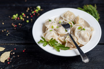 Boiled dumplings with feathers of green onions. In the background are greens, red peppers and bay leaves. On a black wooden background.