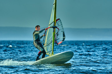A male athlete is interested in windsurfing. He moves on a Sailboard on a large lake on an autumn day.