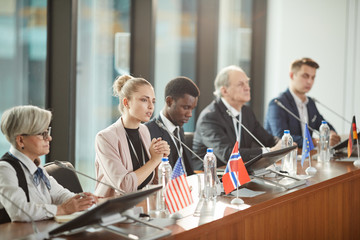 Multiethnic group of political leaders sitting and listening to young woman speaking into the microphone at press conference