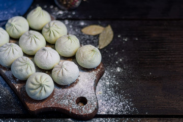 Frozen khinkali on a wooden board, sprinkled with flour. In the background pepper and bay leaf. On a black wooden background.