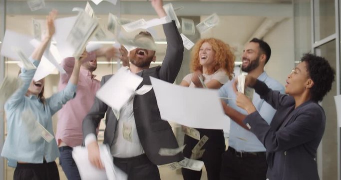 Male and female office workers dancing with money and documents at open space.Excited business team throwing away dollar banknotes and papers in office. Concept of successful business.