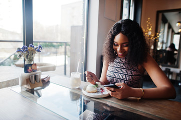 Fototapeta na wymiar Сharming elegant young african american woman with long curly hair wearing jumpsuit sitting at cafe indoor eating dessert and look at mobile phone.