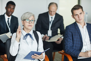 Senior businesswoman with notepad raising her hand while sitting together with her colleagues at seminar