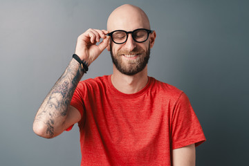 Stylish young bearded man fixing his black framed glasses with a tattooed hand while smiling in...