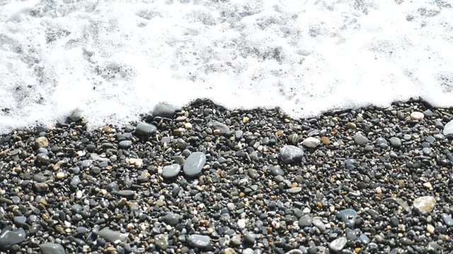 Small gentle waves splashing to pebble beach at sunny day. Ocean tides washing up stony shore. Wet stones sparking from bright sunlight. Beautiful seascape. Travel concept. Slow motion Close up