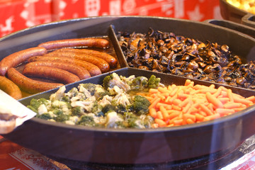 Cauliflower, mushrooms, carrots, sausages fry on a big round pan in a showcase of fast food cafe.