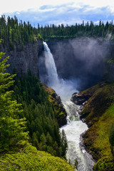Helmcken Waterfall towers above Murtle River with a constant flow of water gushing over the rocky cliff face feeding the dense pine forest it sits within on a partially sunny day in Canada.