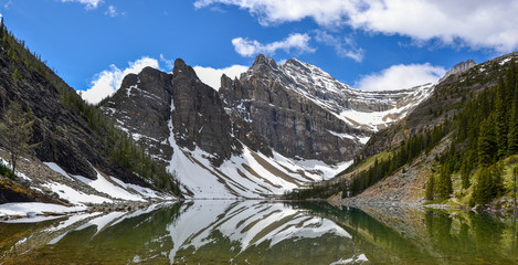 The mirror like reflections on Lake Agnes create beautiful patterns against the mountainous backdrop spotted with snow and pine trees on a sunny and partially cloudy day.