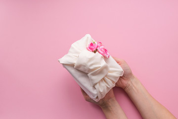 Female hands holding a holiday gift packed fabric in the manner of Furoshiki on a fashionable pink background.
