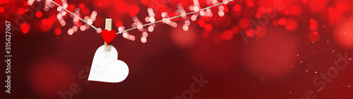 Happy Valentine's Day background banner panorama - White heart hang on wooden clothes pegs with wooden heart and bokeh lights on a string isolated on red texture, with space for text